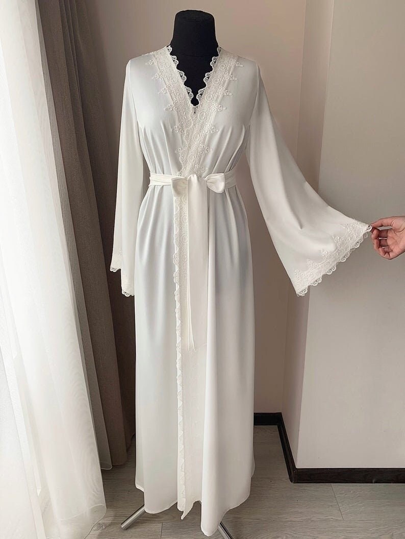Peut inclure: Une robe blanche &agrave; manches longues avec une bordure en dentelle autour du col, des poignets et de la ceinture. La robe a un col en V et une ceinture qui se noue &agrave; la taille.