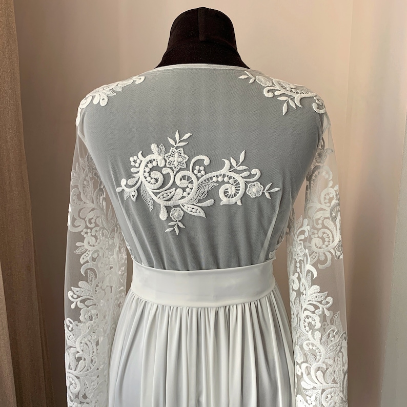 May include: White lace robe with long sleeves and a floral design on the back. The robe has a satin sash and a flowing skirt.