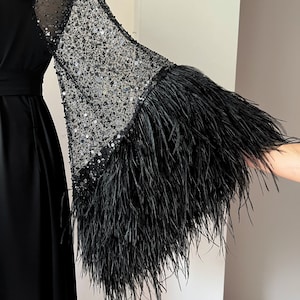 K&ouml;nnte beinhalten: Ein schwarzes Maxikleid mit langen &Auml;rmeln. Die &Auml;rmel sind aus durchsichtigem schwarzen Netzstoff mit Pailletten und einem schwarzen Federnbesatz.