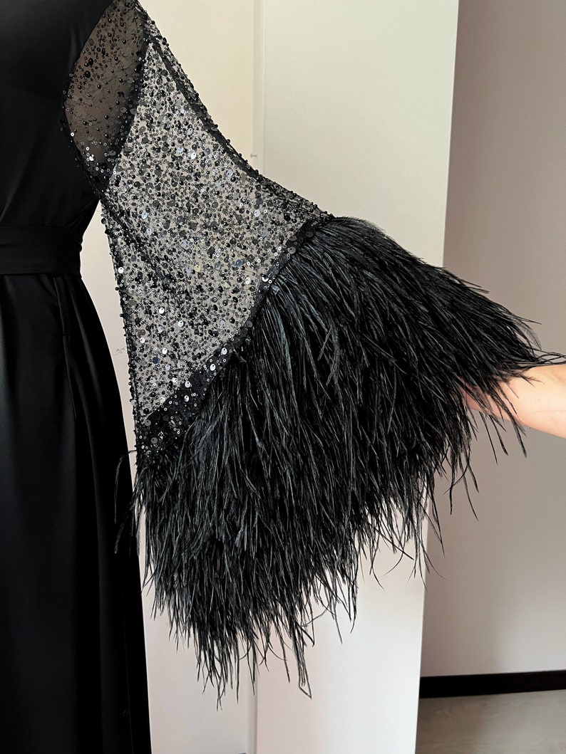 K&ouml;nnte beinhalten: Ein schwarzes Maxikleid mit langen &Auml;rmeln. Die &Auml;rmel sind aus durchsichtigem schwarzen Netzstoff mit Pailletten und einem schwarzen Federnbesatz.