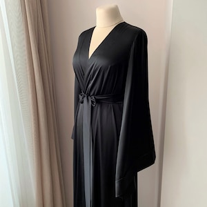 Puede incluir: Una bata larga negra con mangas anchas y un cintur&oacute;n a juego. La bata tiene un escote en V profundo y se muestra en un maniqu&iacute;. La tela parece ser un material suave y sedoso. La bata es un dise&ntilde;o cl&aacute;sico y elegante.