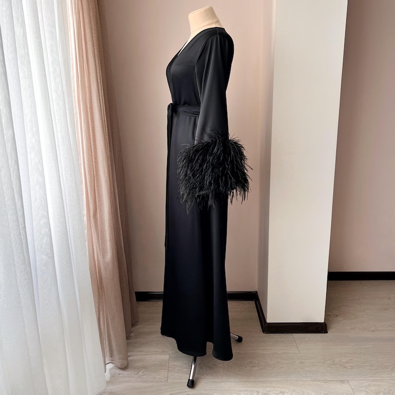 May include: A black satin robe with long sleeves and a feather trim on the cuffs. The robe is tied at the waist with a matching sash.