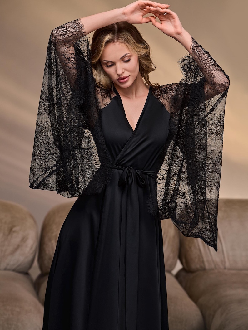 May include: Black robe with a V-neckline and a tie waist. The robe features wide, sheer lace sleeves. The model is wearing the robe with arms raised. The background is a neutral color.