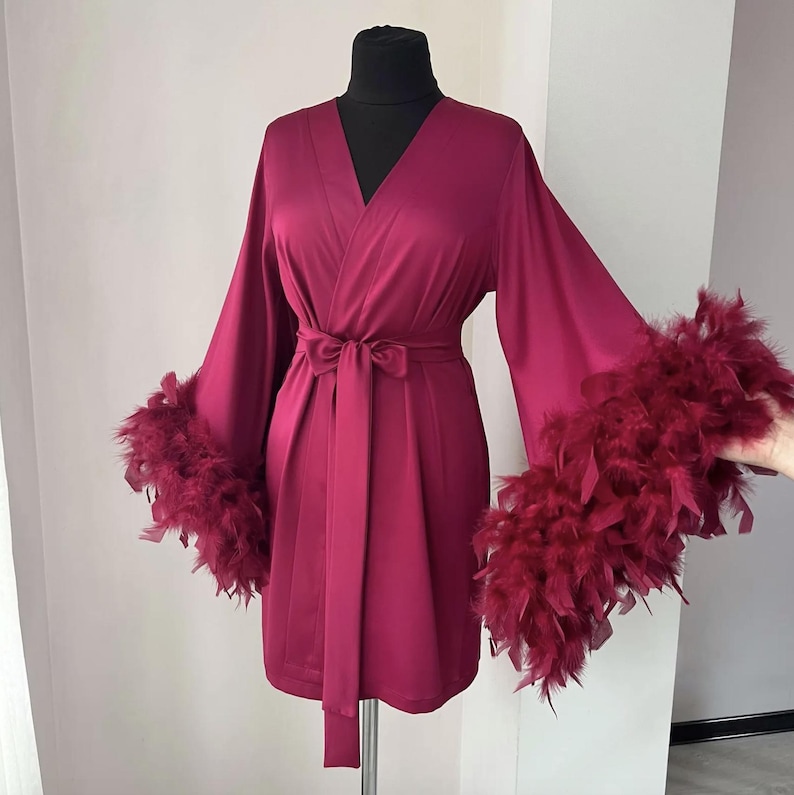 May include: A deep magenta satin robe featuring a wrap-style front with a matching tie belt at the waist and wide, long sleeves finished with thick, matching magenta feather trim at the cuffs, displayed on a black mannequin torso.