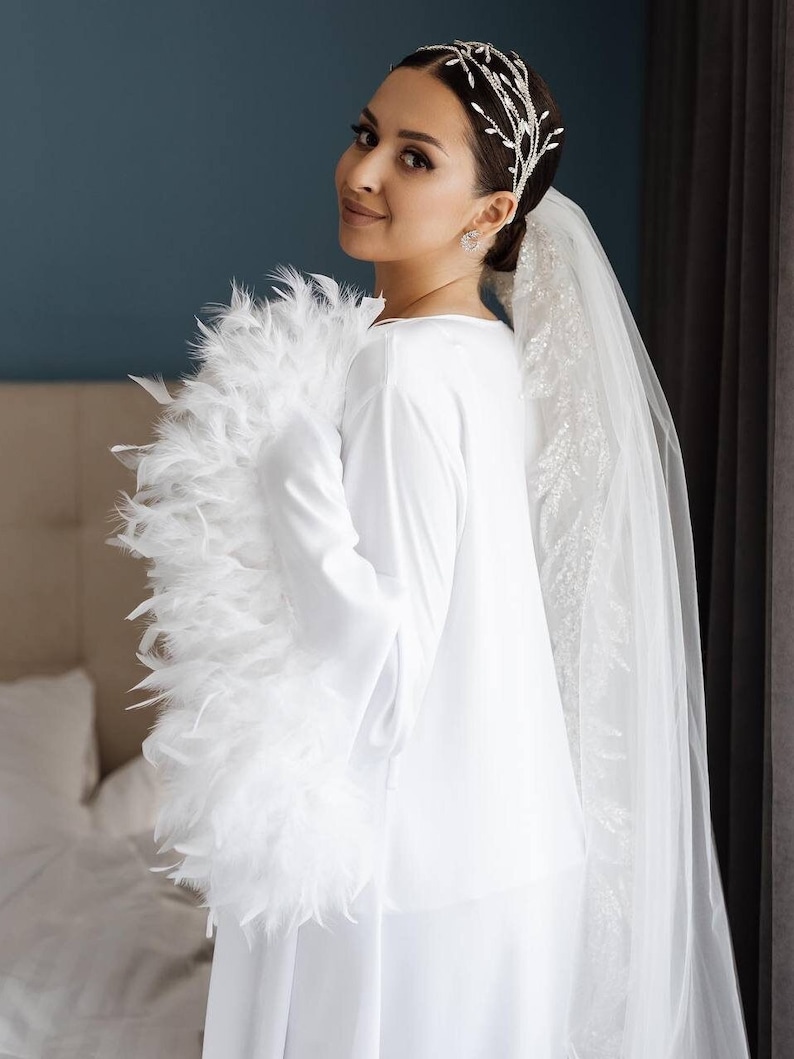 K&ouml;nnte beinhalten: Ein wei&szlig;es Langarmshirt mit volumin&ouml;sem wei&szlig;em Federbesatz an den &Auml;rmeln, kombiniert mit einem transparenten wei&szlig;en Schleier und einem silberfarbenen Kopfschmuck, der mit klaren Kristallen in einem zarten Rankenmuster verziert ist.