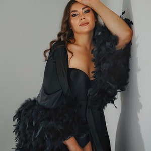 May include: A black satin robe with feather trim. The robe has a long, flowing silhouette and is tied at the waist with a sash. The model is wearing a black bra underneath the robe.