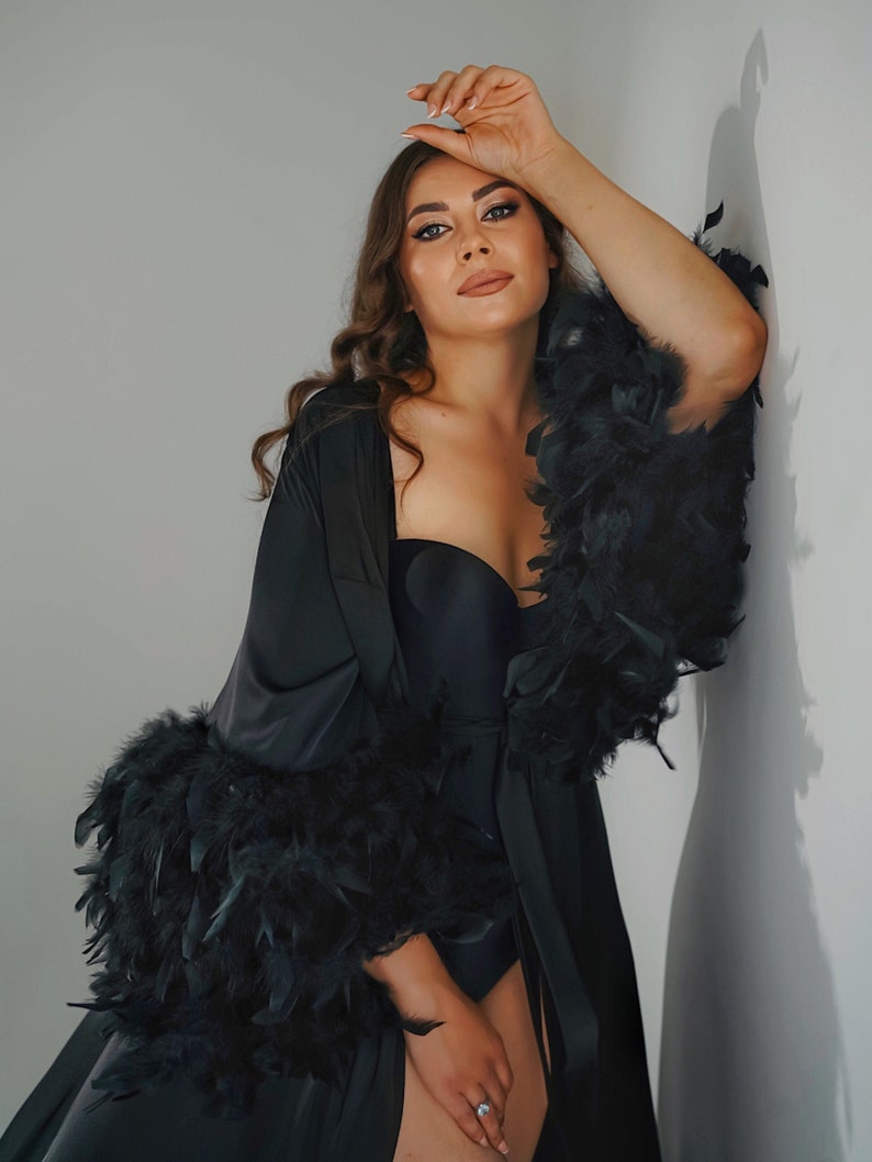 May include: A black satin robe with feather trim. The robe has a long, flowing silhouette and is tied at the waist with a sash. The model is wearing a black bra underneath the robe.