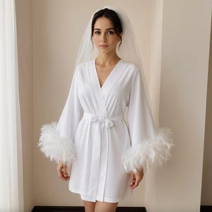 Puede incluir: Bata de satén blanco con cuello en V y cinturón a juego. Las mangas son largas y adornadas con plumas blancas esponjosas. Se lleva un velo blanco transparente en la cabeza. La bata llega a la rodilla.