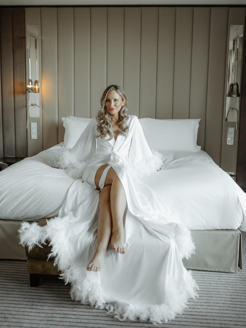 May include: A floor-length white satin robe featuring long, wide sleeves and a hemline trimmed with white feathers. The garment includes a matching fabric tie belt at the waist, creating a wrap-style closure with a deep V-neckline.