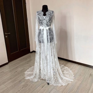 May include: A white lace robe with long sleeves and a flowing train is displayed on a mannequin. The robe features intricate floral patterns and a matching white satin belt tied at the waist. The garment is designed for a special occasion.