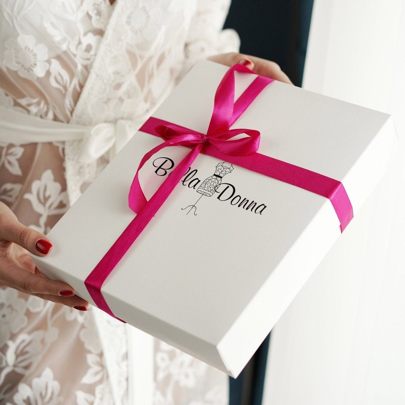 May include: A white rectangular gift box tied with a bright pink ribbon. The box has the words "Bella Donna" printed on it, along with a sketch of a dress form. The box is being held by someone wearing a white lace robe.