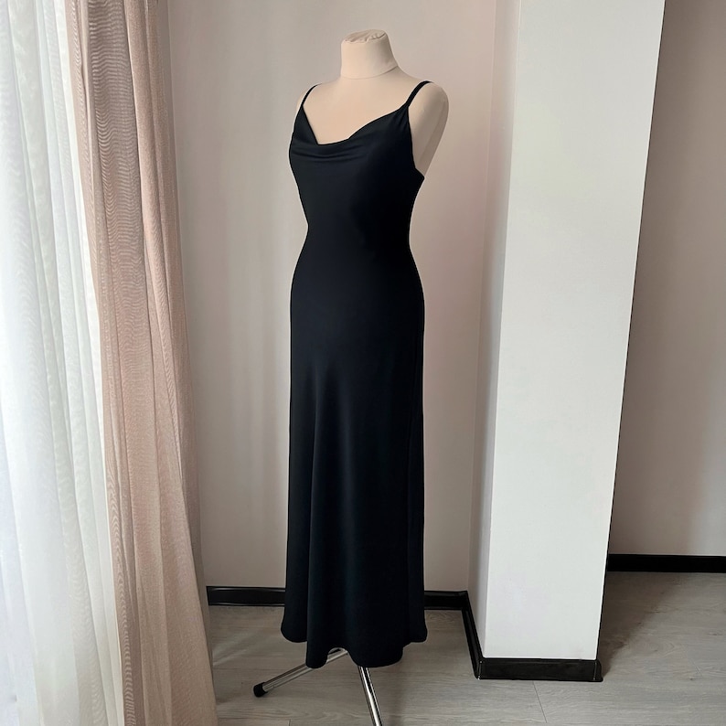K&ouml;nnte beinhalten: Ein schwarzes, &auml;rmelloses Maxikleid mit einem Cowl-Ausschnitt und einem flie&szlig;enden Rock. Das Kleid befindet sich auf einer Schaufensterpuppe und wird vor einer wei&szlig;en Wand pr&auml;sentiert.