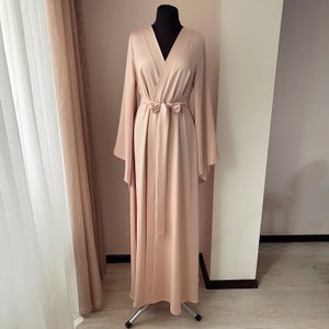 May include: A long, blush-colored satin robe with a V-neckline and wide sleeves. The robe has a sash tie at the waist.