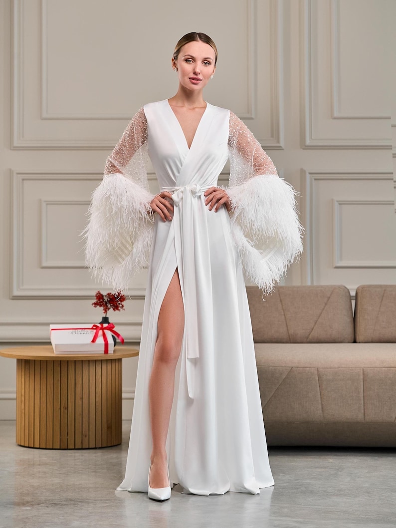 A bride poses confidently in a white satin bridal robe with sheer beaded sleeves and dramatic ostrich feather cuffs. The robe features a deep V-neckline, a waist-cinching belt, and a high slit, creating a luxurious and elegant silhouette.