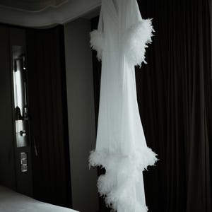 May include: A long white robe featuring a V-neckline and long sleeves, with thick white feather trim detailing along the sleeve cuffs and the bottom hemline. The garment hangs vertically, displaying a smooth fabric texture and a full-length silhouette.