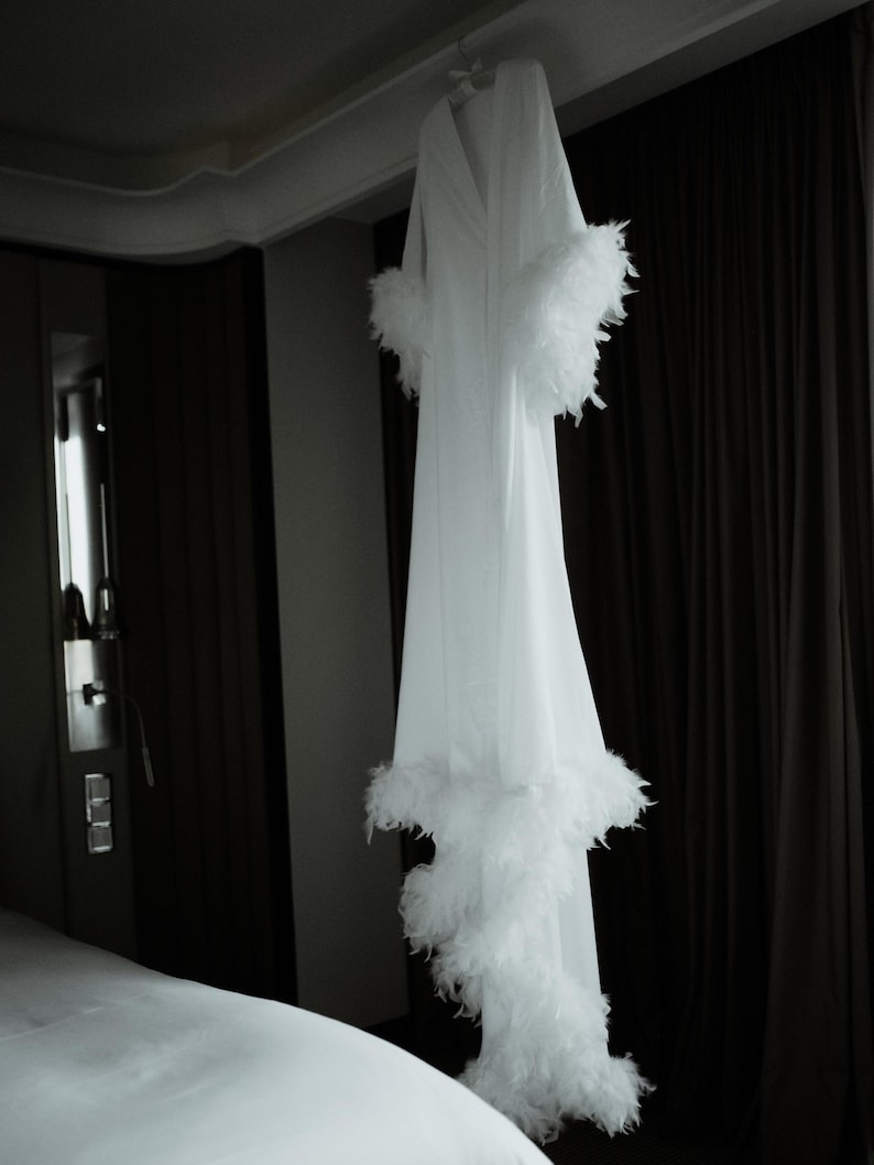 May include: A long white robe featuring a V-neckline and long sleeves, with thick white feather trim detailing along the sleeve cuffs and the bottom hemline. The garment hangs vertically, displaying a smooth fabric texture and a full-length silhouette.