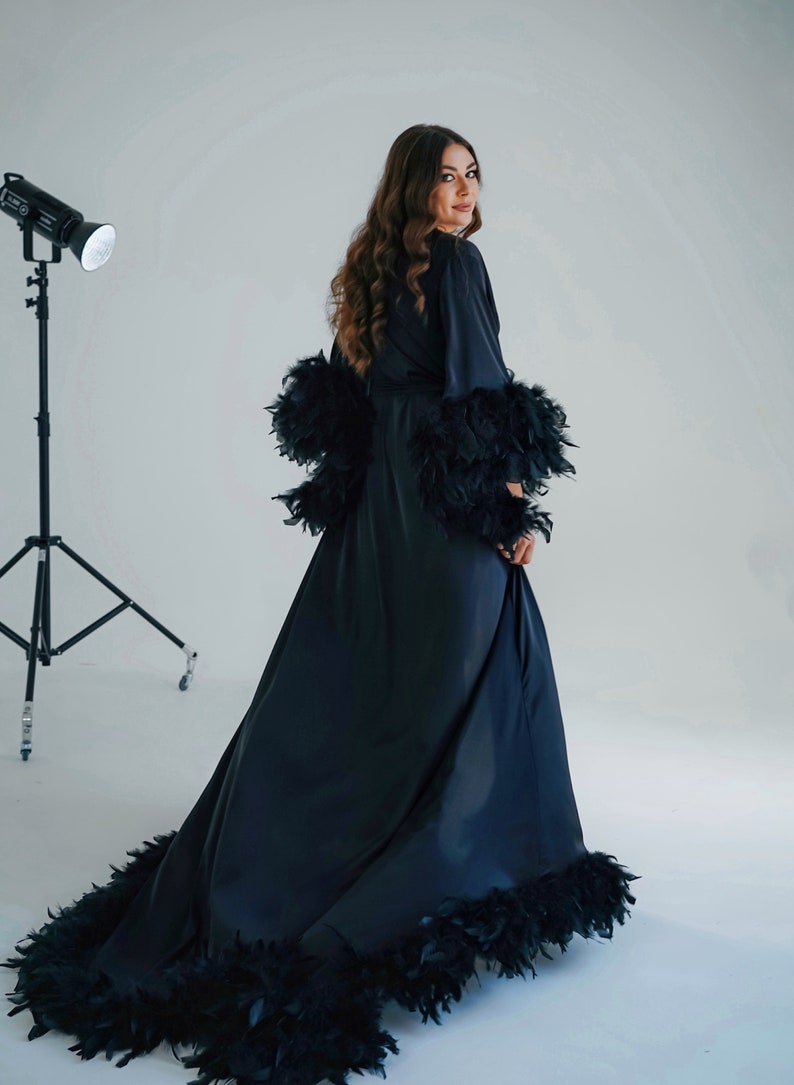 May include: A long black satin robe with feather trim around the cuffs, hem, and neckline. The robe is worn by a person with long brown hair.