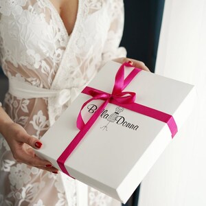 Peut inclure: Une bo&icirc;te cadeau blanche avec un ruban rose et le texte "Bella Donna" imprim&eacute; sur le dessus. La bo&icirc;te est tenue par une personne portant une robe en dentelle blanche.