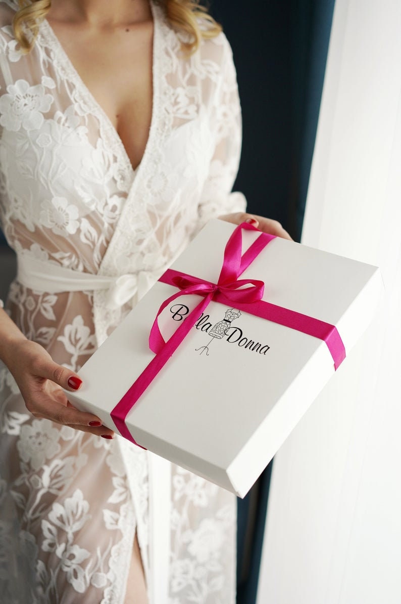 Peut inclure: Une bo&icirc;te cadeau blanche avec un ruban rose et le texte "Bella Donna" imprim&eacute; sur le dessus. La bo&icirc;te est tenue par une personne portant une robe en dentelle blanche.