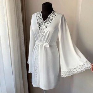 May include: A white satin robe with wide sleeves and a lace trim. The robe has a V-neckline with lace detailing and a matching tie belt. The robe is displayed on a black mannequin.