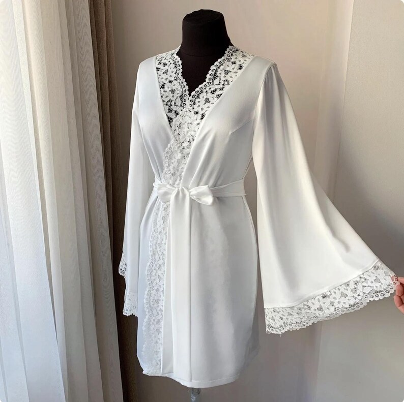 May include: A white satin robe with wide sleeves and a lace trim. The robe has a V-neckline with lace detailing and a matching tie belt. The robe is displayed on a black mannequin.