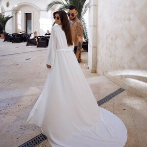 May include: A woman in a white robe with long sleeves and a long train walks away from the camera. She is wearing sunglasses and has long brown hair. A man in swim trunks is walking behind her.