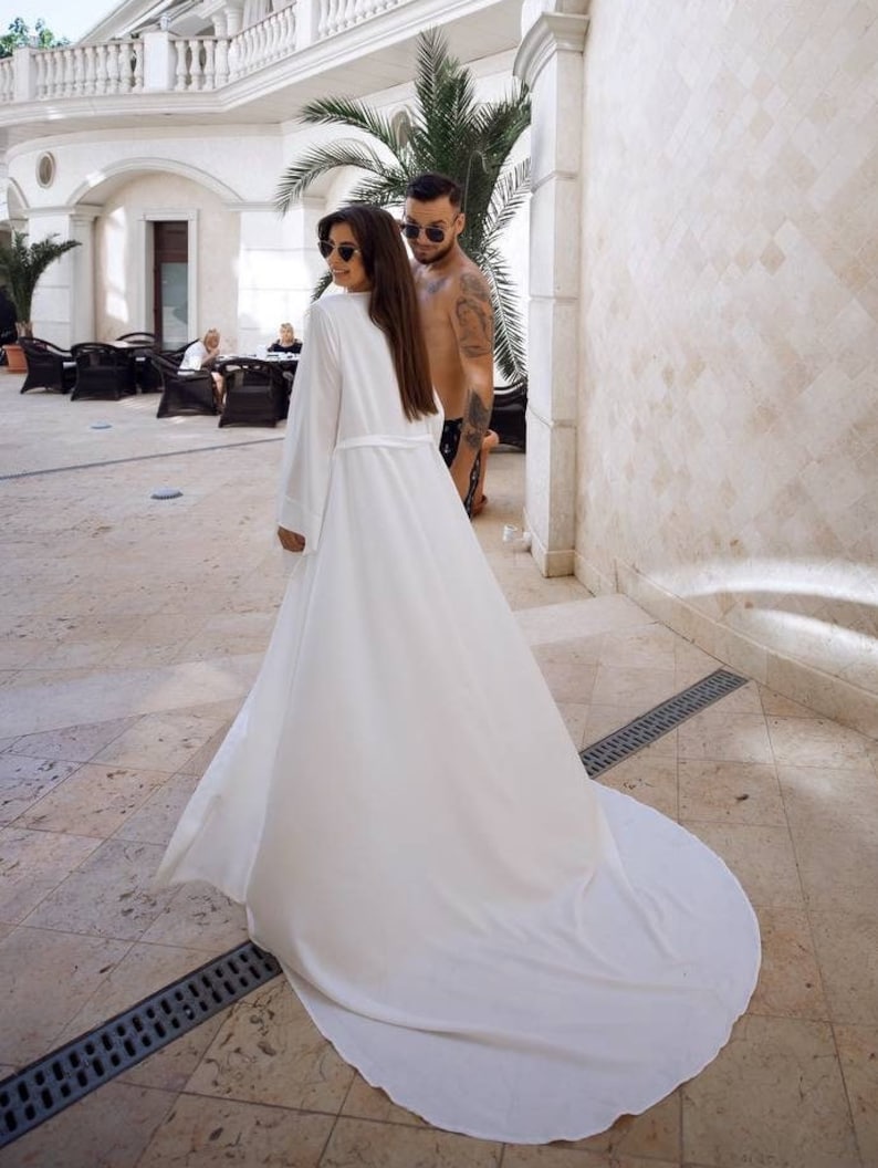 May include: A woman in a white robe with long sleeves and a long train walks away from the camera. She is wearing sunglasses and has long brown hair. A man in swim trunks is walking behind her.