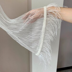 May include: A white sheer mesh sleeve with a flared bell shape, featuring a wide band of white beaded trim at the cuff and a long, flowing fringe of white feathers extending from the edge of the beaded trim, creating a textured appearance.