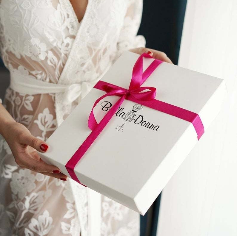 May include: A white gift box with a pink ribbon and the text "Bella Donna" printed on the top. The box is being held by a person wearing a white lace robe.