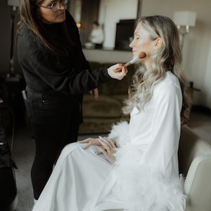 May include: A white satin robe featuring long sleeves with thick white feather trim at the cuffs. The robe has a wrap-style front with a matching tie belt, creating a smooth, flowing silhouette that reaches the floor while worn in a seated position.