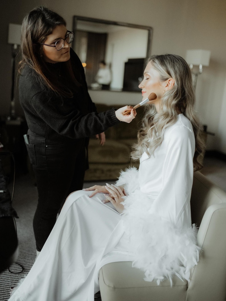 May include: A white satin robe featuring long sleeves with thick white feather trim at the cuffs. The robe has a wrap-style front with a matching tie belt, creating a smooth, flowing silhouette that reaches the floor while worn in a seated position.