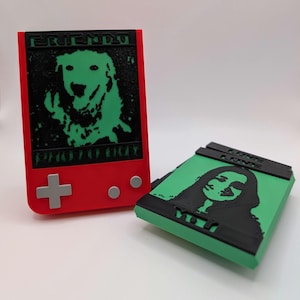 May include: Two 3D printed handheld game consoles with different designs. The first console is red with a green image of a dog and the text "FRIENDLY PHOTO BOOT". The second console is green with a black image of a person and the text "LOVE LOVE YOU".