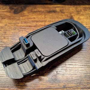 Puede incluir: Un organizador de electrónica de plástico negro impreso en 3D. Tiene un compartimento para un teléfono, una ranura para un cable USB y un espacio para tarjetas SD.