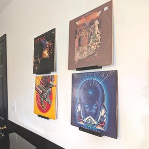 May include: Four vinyl records are displayed on a white wall using black metal record holders. The records are "Styx - Pieces of Eight", "Journey - Frontiers", "Styx -  The Grand Illusion", and "The Eagles -  Hotel California".