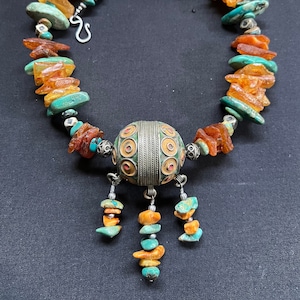 May include: A silver and turquoise necklace with a large, round pendant. The pendant is decorated with intricate silverwork and red and orange accents. The necklace is made of amber and turquoise beads.