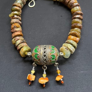 May include: A necklace made of green, brown and yellow stones with a silver pendant. The pendant is round and has a green and red enamel design.