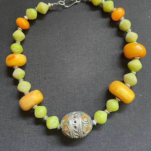 Puede incluir: Collar de cuentas con cuentas verdes, naranjas y amarillas. La cuenta central es plateada, con detalles de esmalte de colores. Las cuentas están ensartadas en una cadena plateada con un cierre de gancho. Longitud aproximada 45 cm.