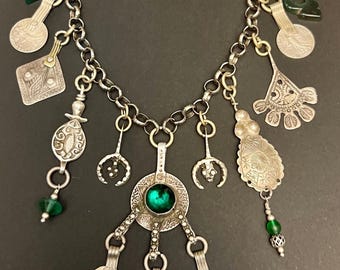 May include: Silver-toned necklace with various charms, including coins, geometric shapes, and green beads. The necklace features a central pendant with a large, round, green gemstone. The chain is made of interconnected links and has a hook clasp.