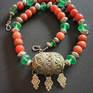 May include: A silver necklace with a large, ornate pendant. The pendant is made of silver and has a green and silver design. The necklace is made of coral and green glass beads.