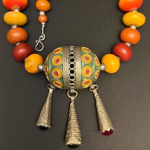 May include: A silver and enamel necklace with a large, round pendant. The pendant is decorated with green, yellow, and red circles. The necklace is made of amber beads in shades of orange, red, and yellow.