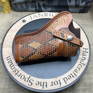 May include: A handcrafted leather holster with a snakeskin pattern. The holster is brown and tan with a black interior. The holster is displayed on a wooden stand with the text "Handcrafted for the Sportsman".