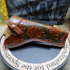 May include: Hand-tooled leather holster with floral and leaf designs in shades of brown, green, and orange. The holster is displayed on a black surface with a wooden base that reads "Handcrafted for the Sportsman."