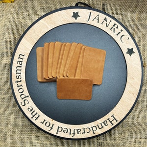 May include: A stack of 10 brown leather patches with the text "Handcrafted for the Sportsman" and "Janric" on a black and wood background.