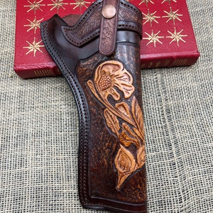 May include: A handcrafted brown leather holster with a floral design. The holster features a detailed embossed flower and leaf pattern, with a contrasting darker brown trim and stitching. A copper-colored button secures the belt loop.