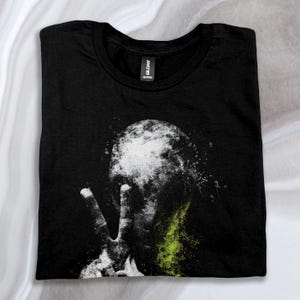 May include: Black t-shirt featuring an abstract design of a person making a peace sign in white and green.