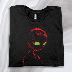 May include: Black t-shirt featuring a red and green alien graphic. The alien has large eyes and a faint, ghostly appearance.