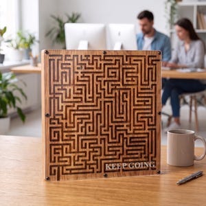 Op de afbeelding: Een vierkante houten doolhofpuzzel met een complex gegraveerd padontwerp achter een doorzichtige acrylplaat. In de rechterbenedenhoek van het houten frame staat de tekst KEEP GOING in witte hoofdletters, vastgezet met vier metalen schroeven.