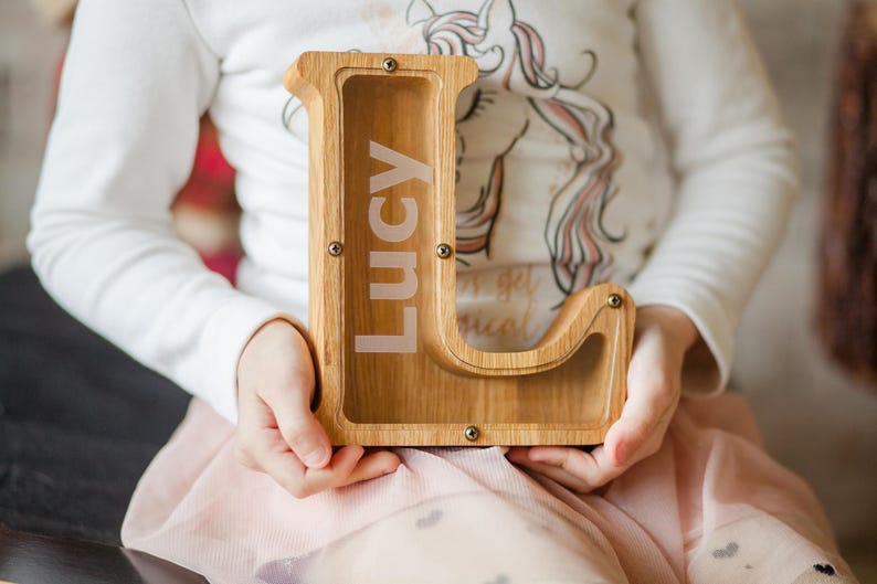 A person is holding a wooden letter bank with the name "Lucy" written on it.