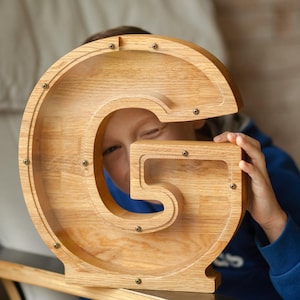 Può includere: Un salvadanaio in legno color marrone chiaro a forma di lettera maiuscola G, dotato di un pannello frontale in acrilico trasparente fissato con piccole viti metalliche. Il contenitore a forma di lettera presenta una venatura del legno visibile.