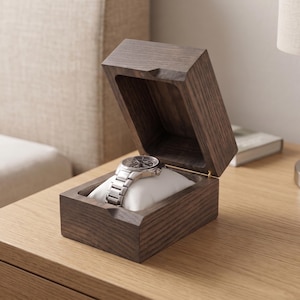 May include: A rectangular dark brown wooden watch box with a hinged lid, featuring a soft white cushion inside that holds a silver-toned metal wristwatch with a link bracelet. The box has a visible wood grain texture and a simple, clean design.